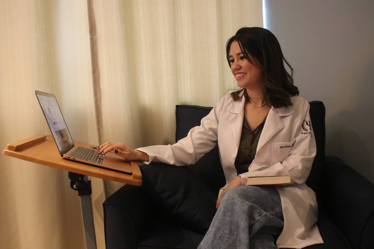Ana Neves atendendo online pelo notebook apoiado em suporte de madeira no consultório